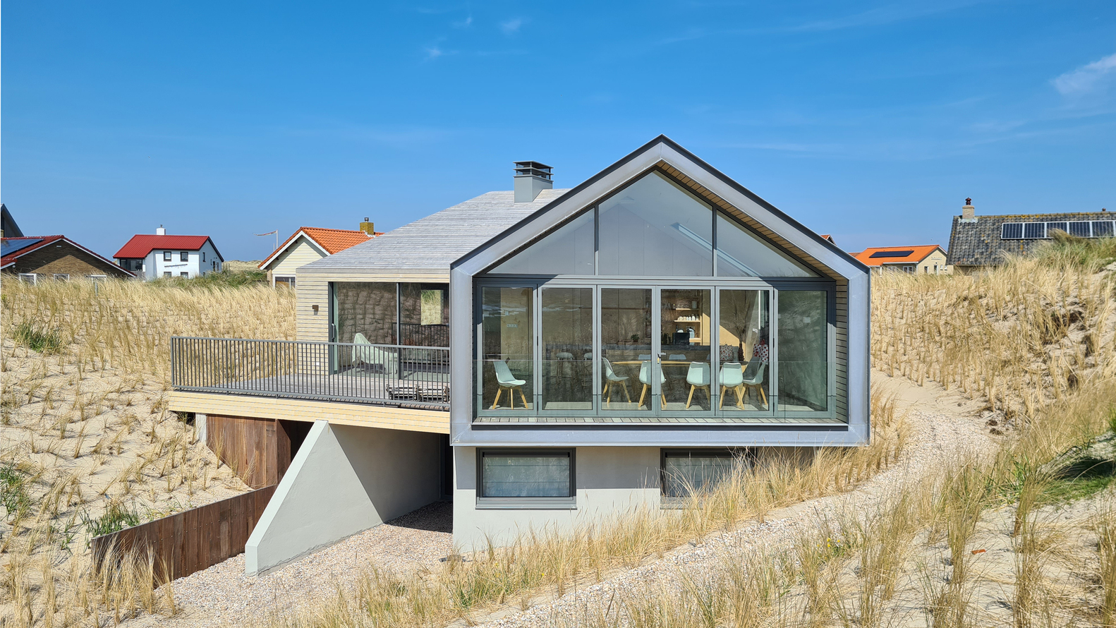 Gabled glazed facade with timber deck and metal railing embedded in dune vegetation between neighboring houses