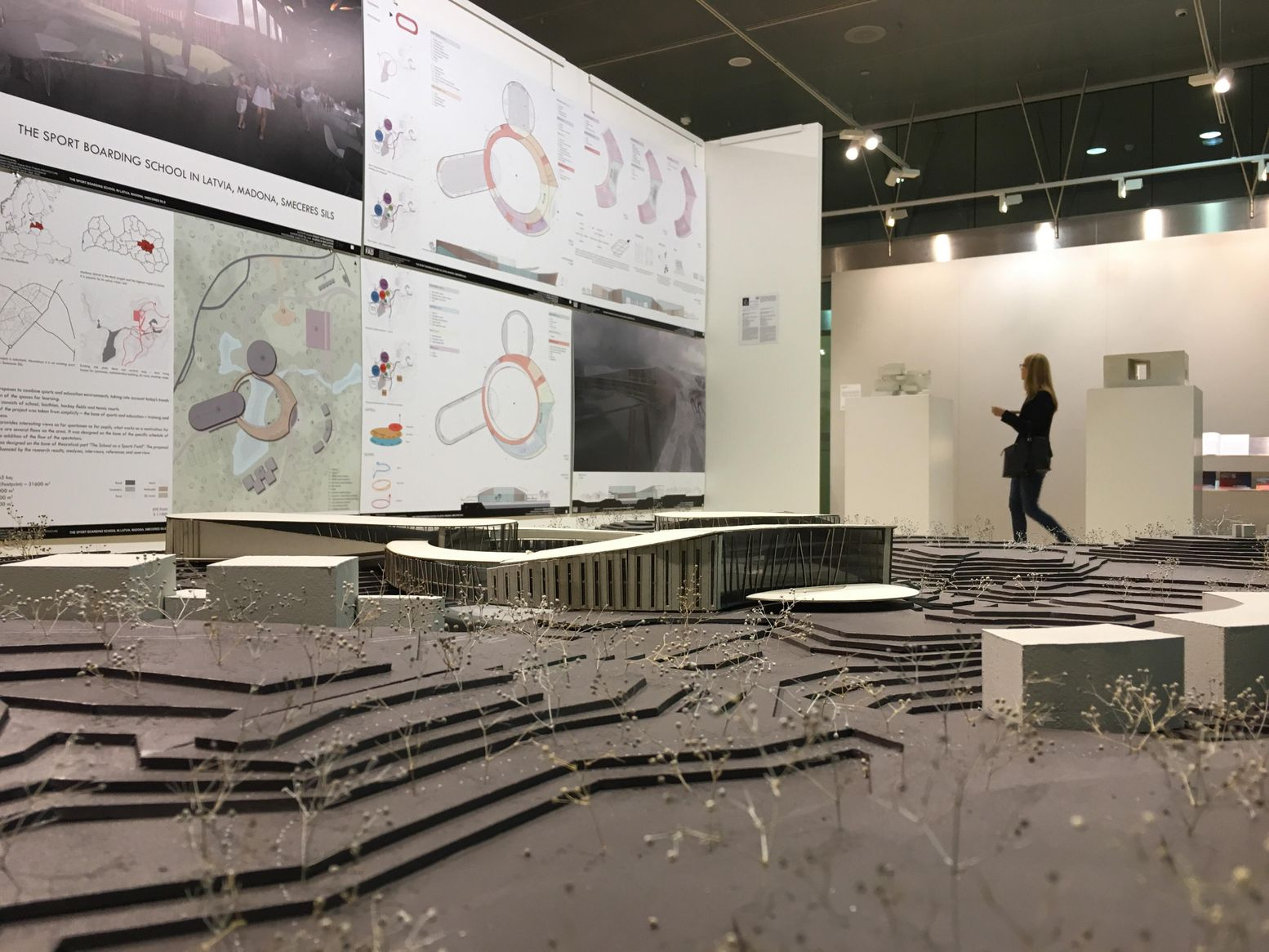Low view of a site model with stepped contour layers, a visitor walking past exhibition boards of plans behind it