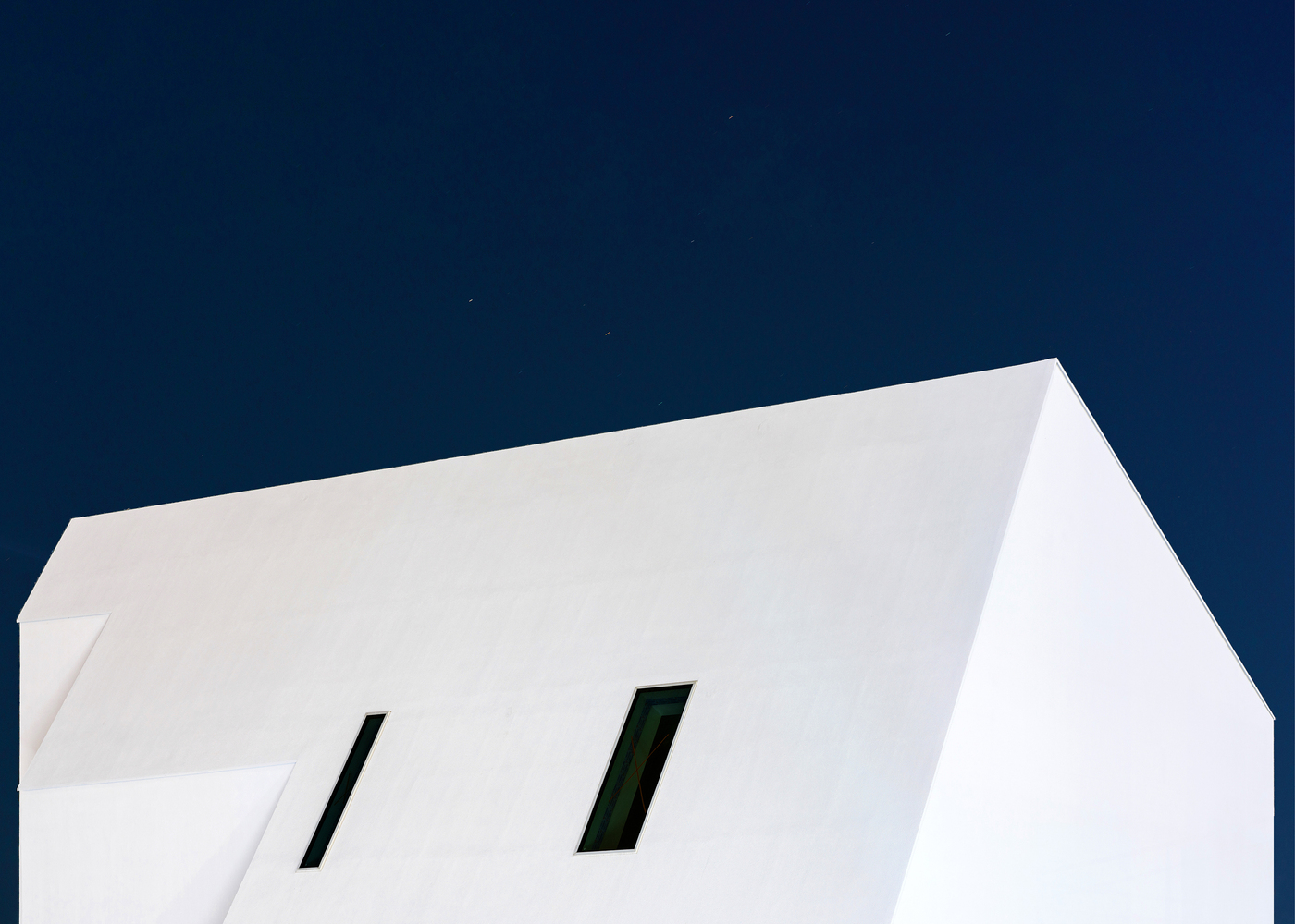 Upward view of white facade with two angled window openings under a deep blue evening sky