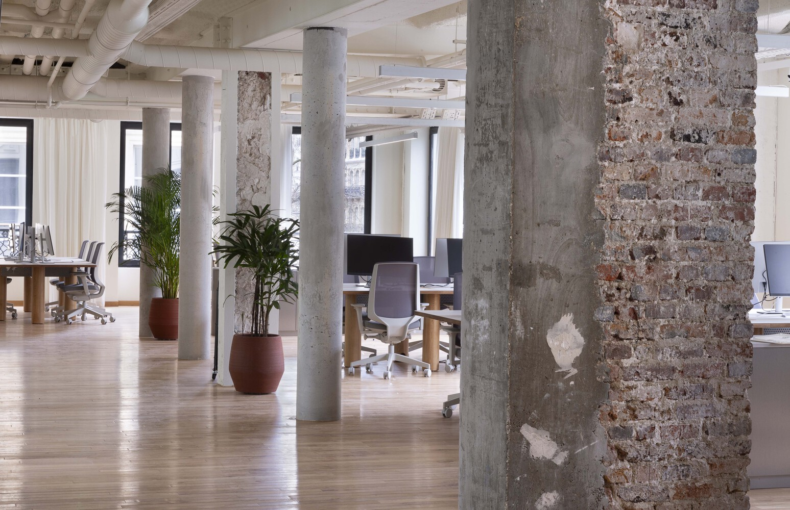 After: open-plan office with exposed concrete and brick columns, oak floors, workstations, plants
