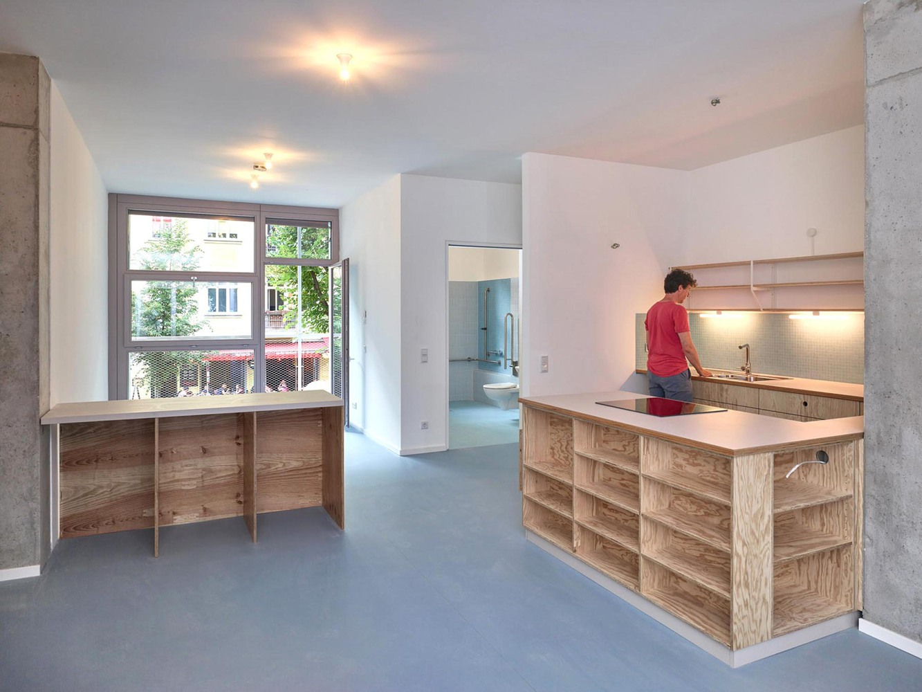 Open kitchen and living area with plywood cabinetry, grey flooring, and exposed concrete columns