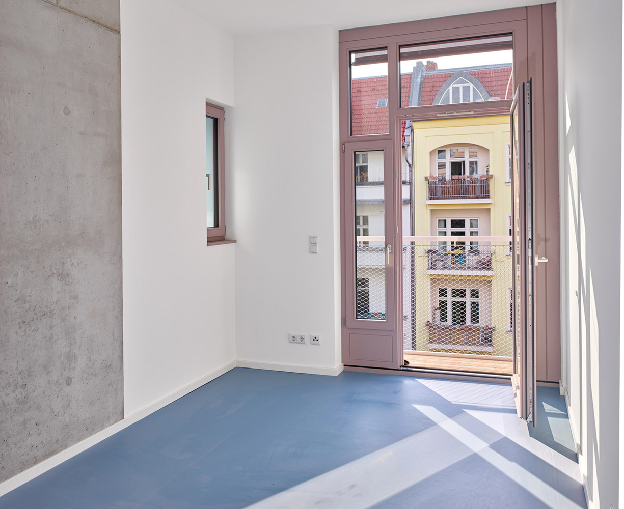 Room with blue flooring, exposed concrete wall, and pink-framed doors opening to balcony with metal mesh railing