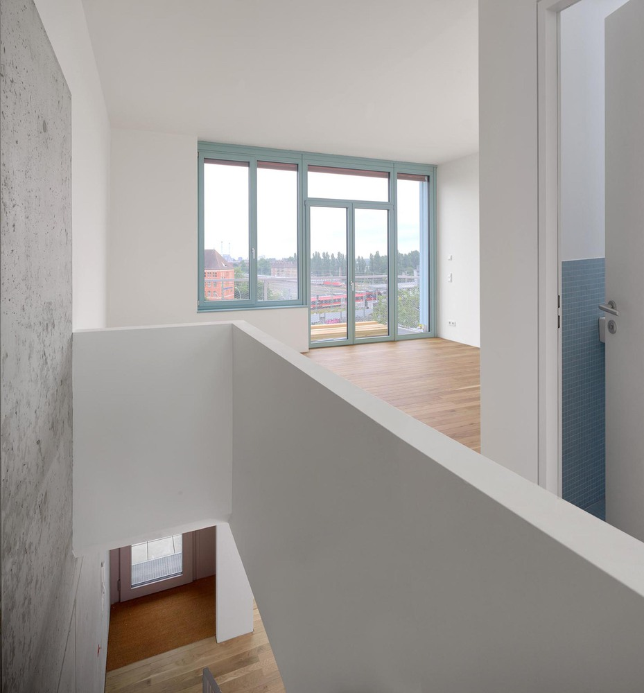 Double-height interior void with white walls, timber flooring, and teal-framed windows overlooking rooftops