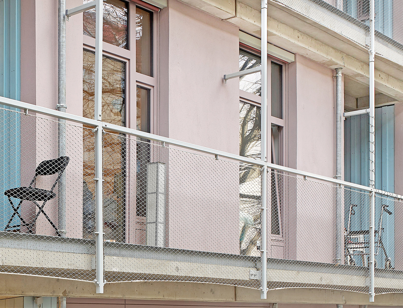 Narrow balcony with white mesh railing, folding chair, and pale pink facade with teal window frames