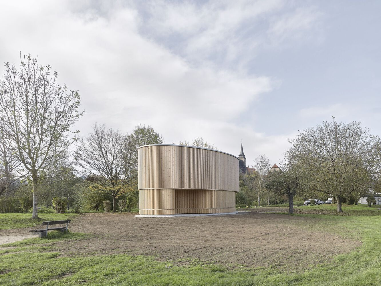 Cylindrical timber pavilion on a park field with church spire visible through bare trees