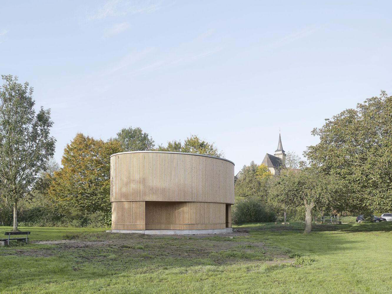 Cylindrical timber-clad volume set in mown grass with church tower beyond autumn tree canopy