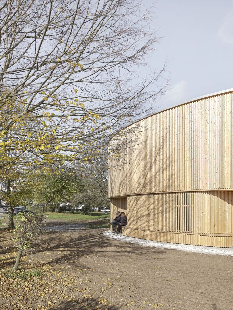 Two people seated on the exterior step beneath the cantilevered upper volume of the timber pavilion
