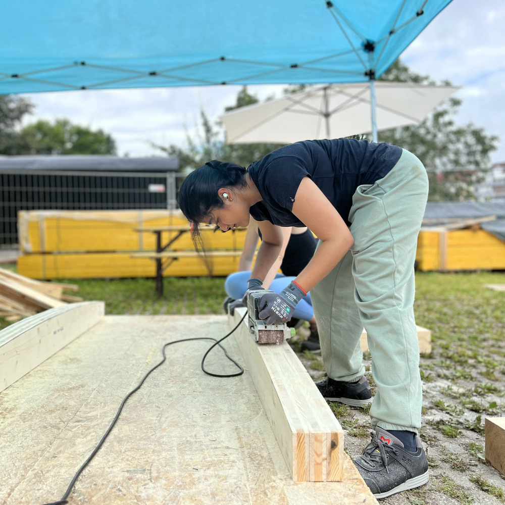 Builder using a power sander on a timber beam under a blue canopy in an outdoor workspace