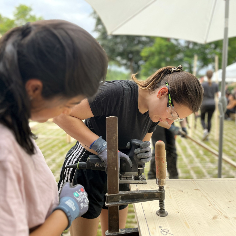 Two builders operating a jigsaw on a plywood sheet clamped to a workbench outdoors