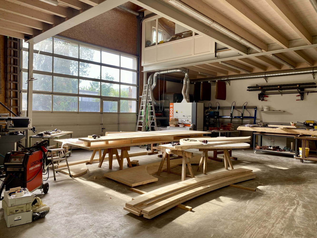 Workshop interior with timber beams on sawhorses beneath exposed ceiling joists and a large gridded window