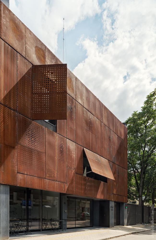 Weathered corten steel facade with perforated panels and hinged shutters beneath a cloudy summer sky