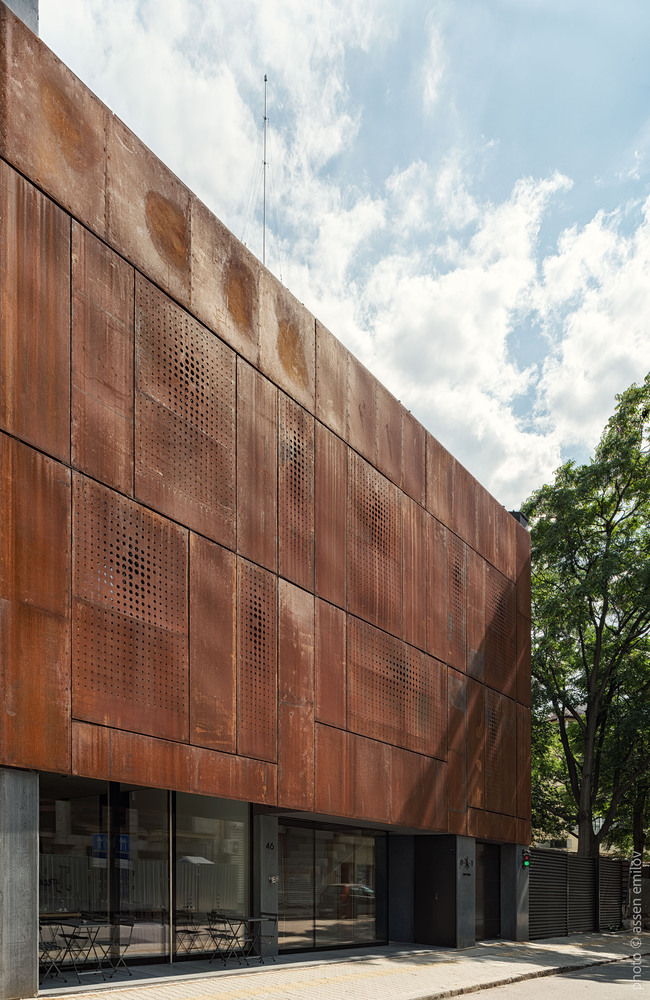 Weathered corten steel facade with perforated panels above glazed ground floor entrance and adjacent tree canopy