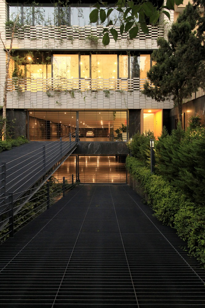 Entry bridge at dusk: black steel footbridge over a reflecting water channel, warm light from the lobby beyond, plants framing the approach