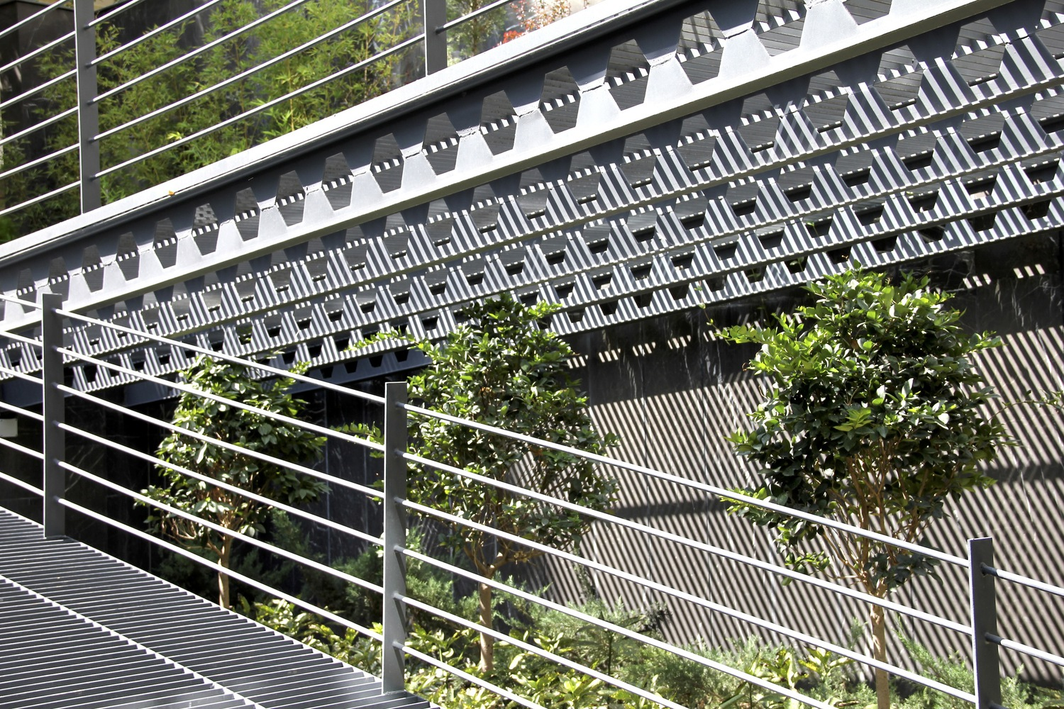 Perforated screen detail: chevron-patterned black steel balcony panel, horizontal cable balustrade, tree foliage in the background