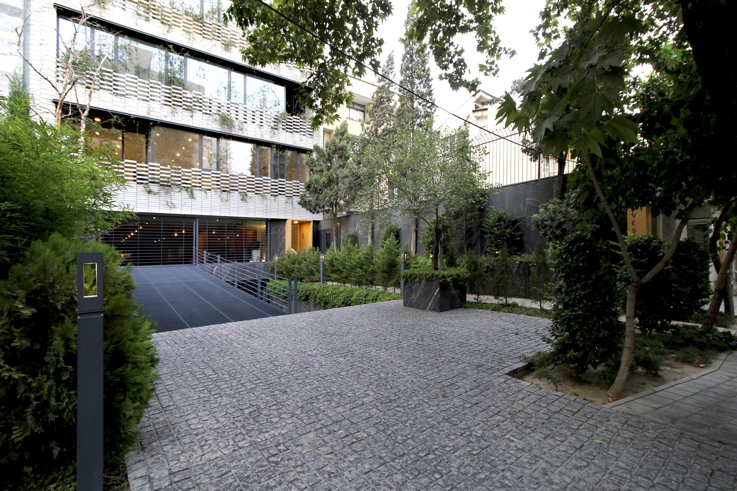 Ground-level entry: stone-paved forecourt with mature trees, building facade beyond with warm lobby light, planted edges, dusk sky