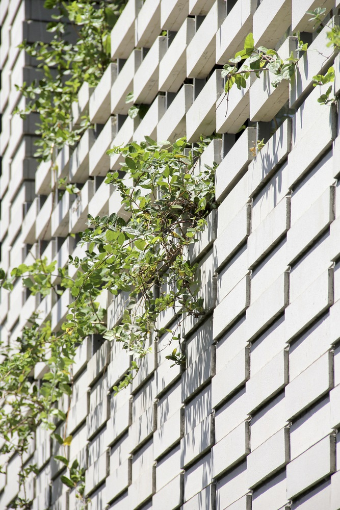 Facade close-up: white perforated metal screens in a chevron pattern, vines hanging from upper balconies, narrow windows between panels