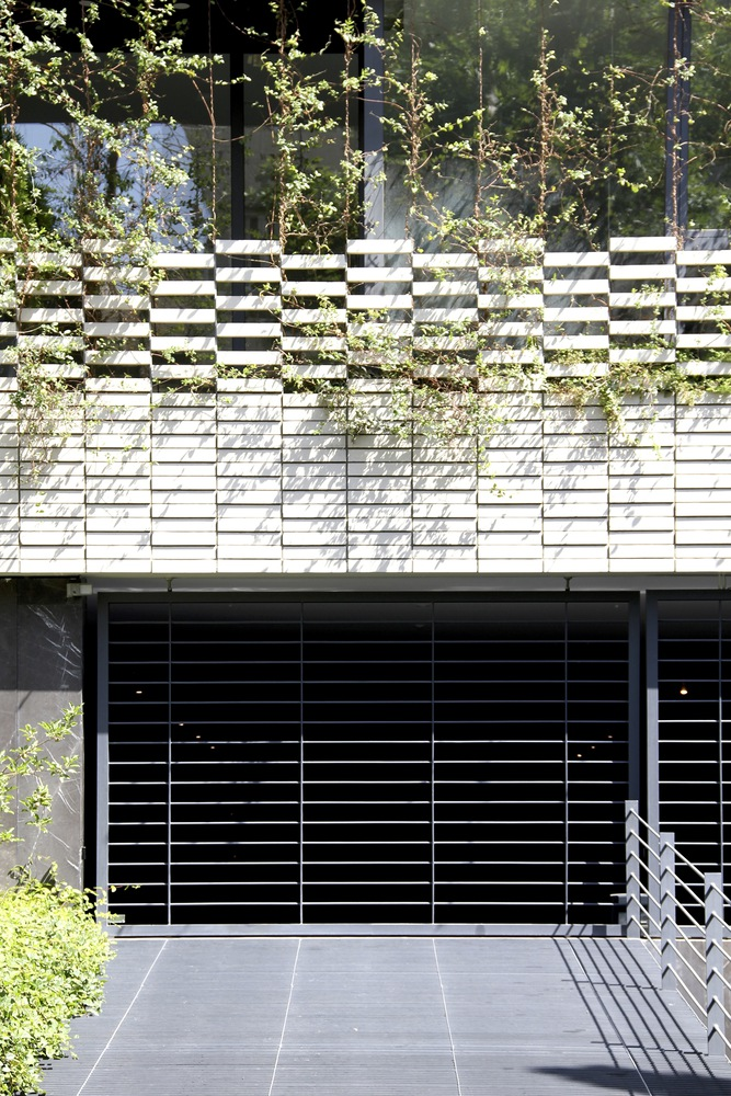 Lower facade: white chequered cladding and perforated steel grille at the basement vent, planters with vines cascading, stone-paved forecourt