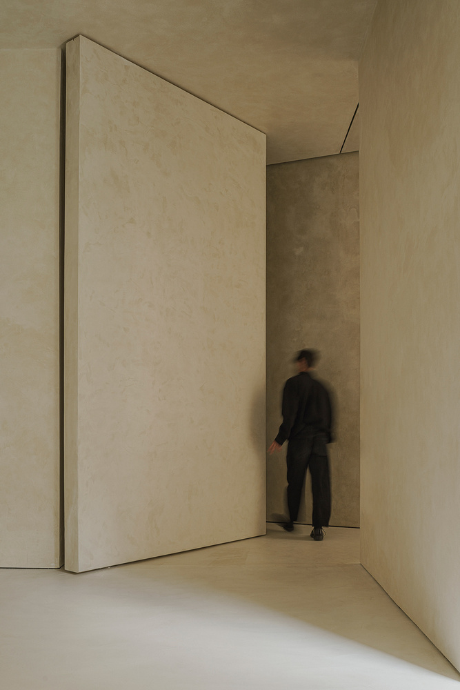 Tall pivoting plaster-clad door with a person walking through the narrow passage in soft light