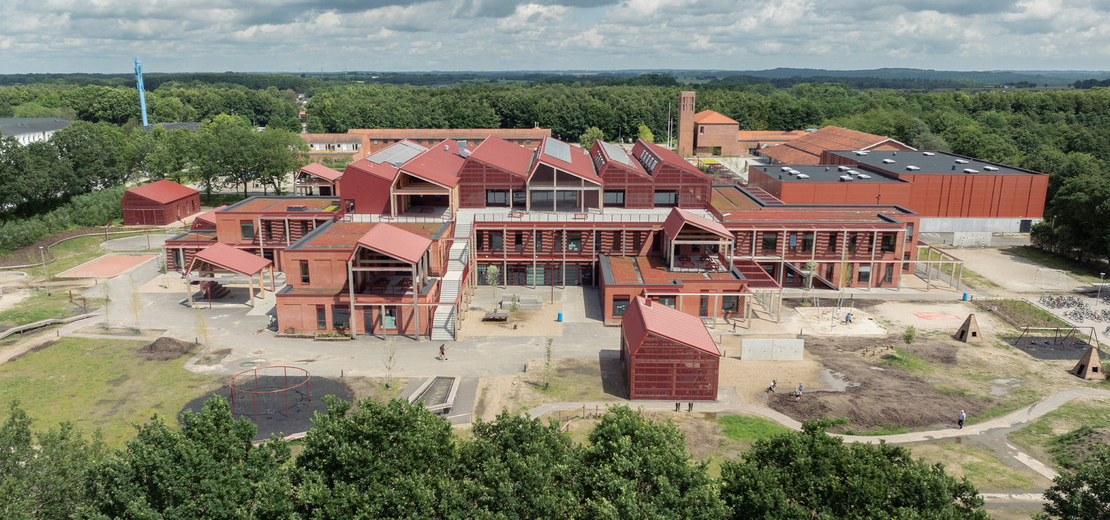 Playground approach: red brick volumes with pitched roofs, external stairs, timber colonnade, timber play logs in foreground