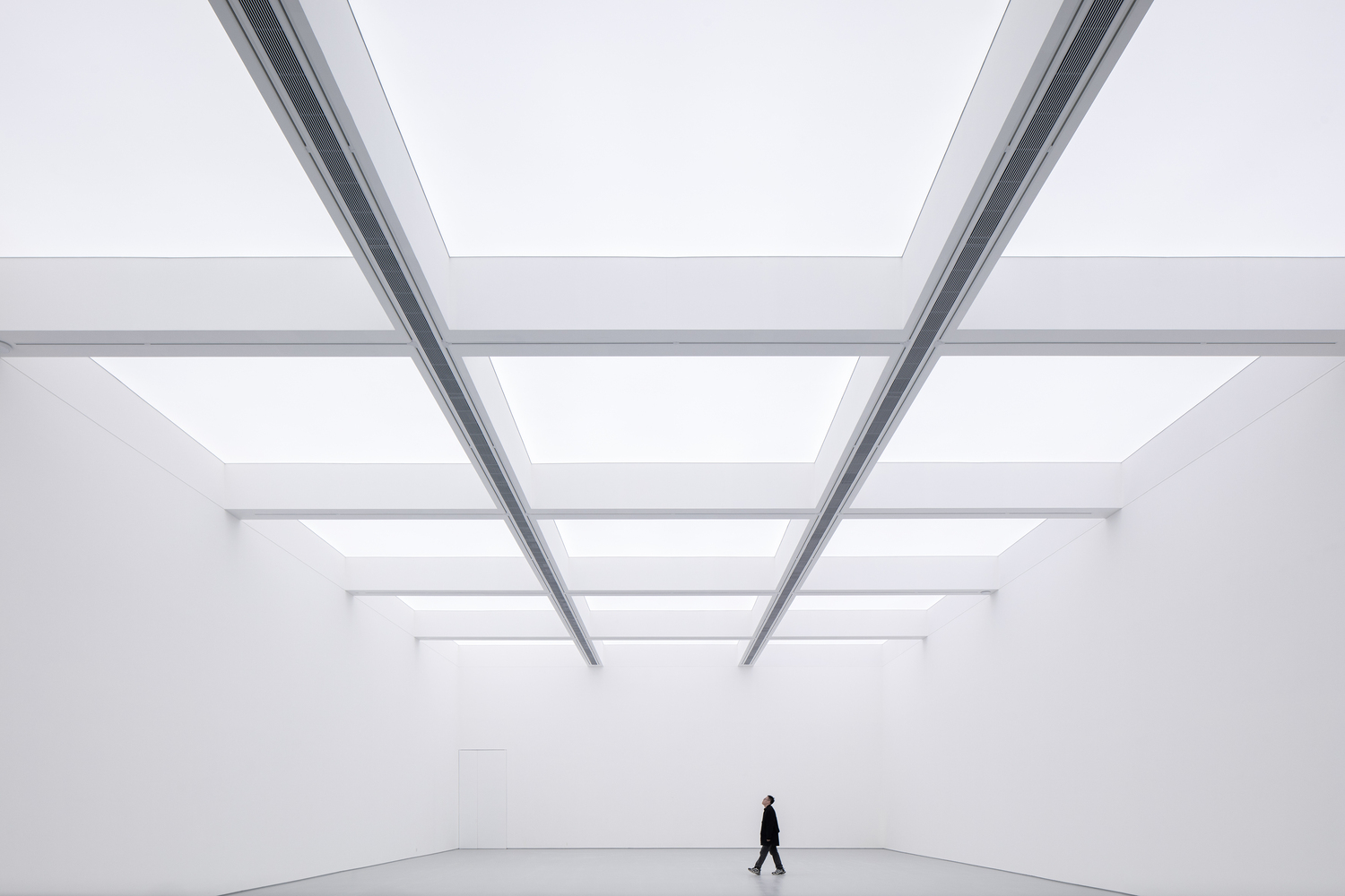 Main hall with figure: person walking across the vast white room under a skylight grid, steel beams converging above