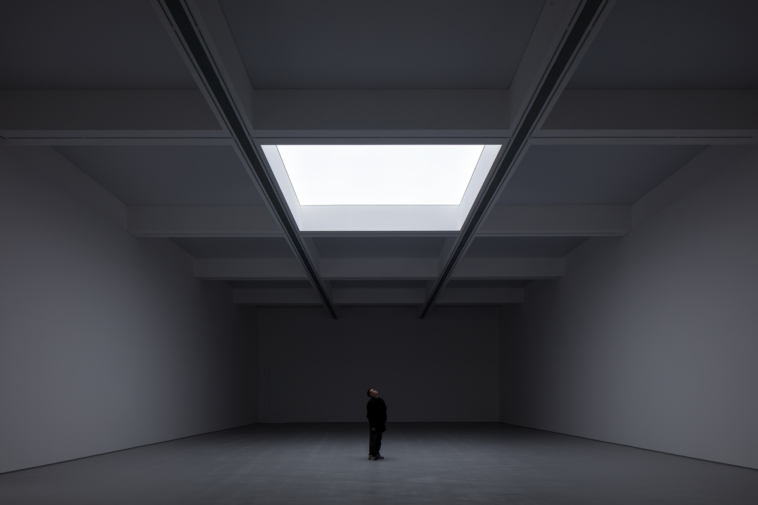 Darkened hall: single central skylight illuminating a dark exhibition space, figure standing below