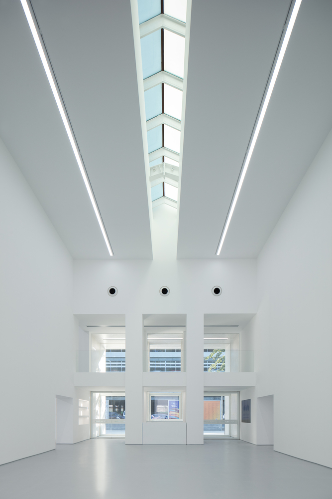 Lobby interior: tall white volume, skylight strip running the length of the ceiling, three windows to the street at the far end