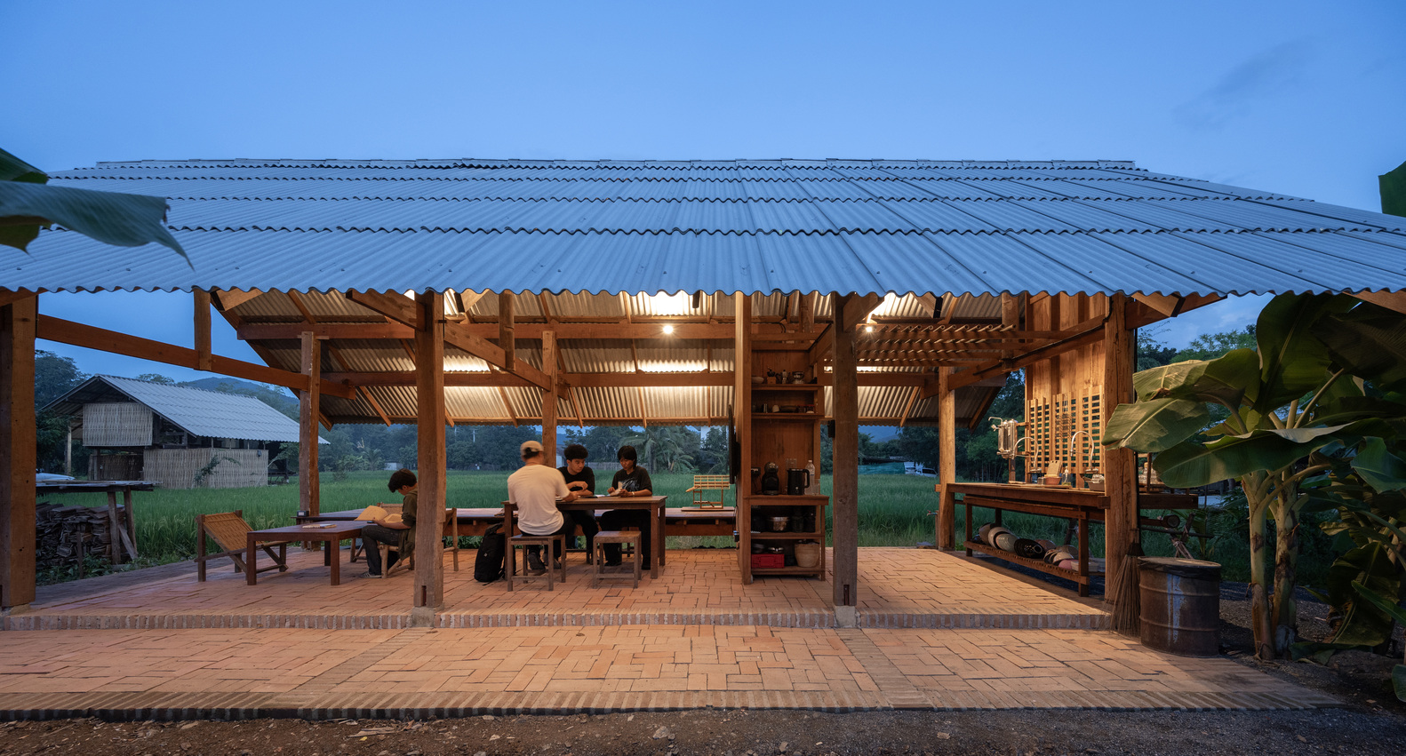 Corrugated metal roof sheltering timber frame structure with people seated at tables under evening light