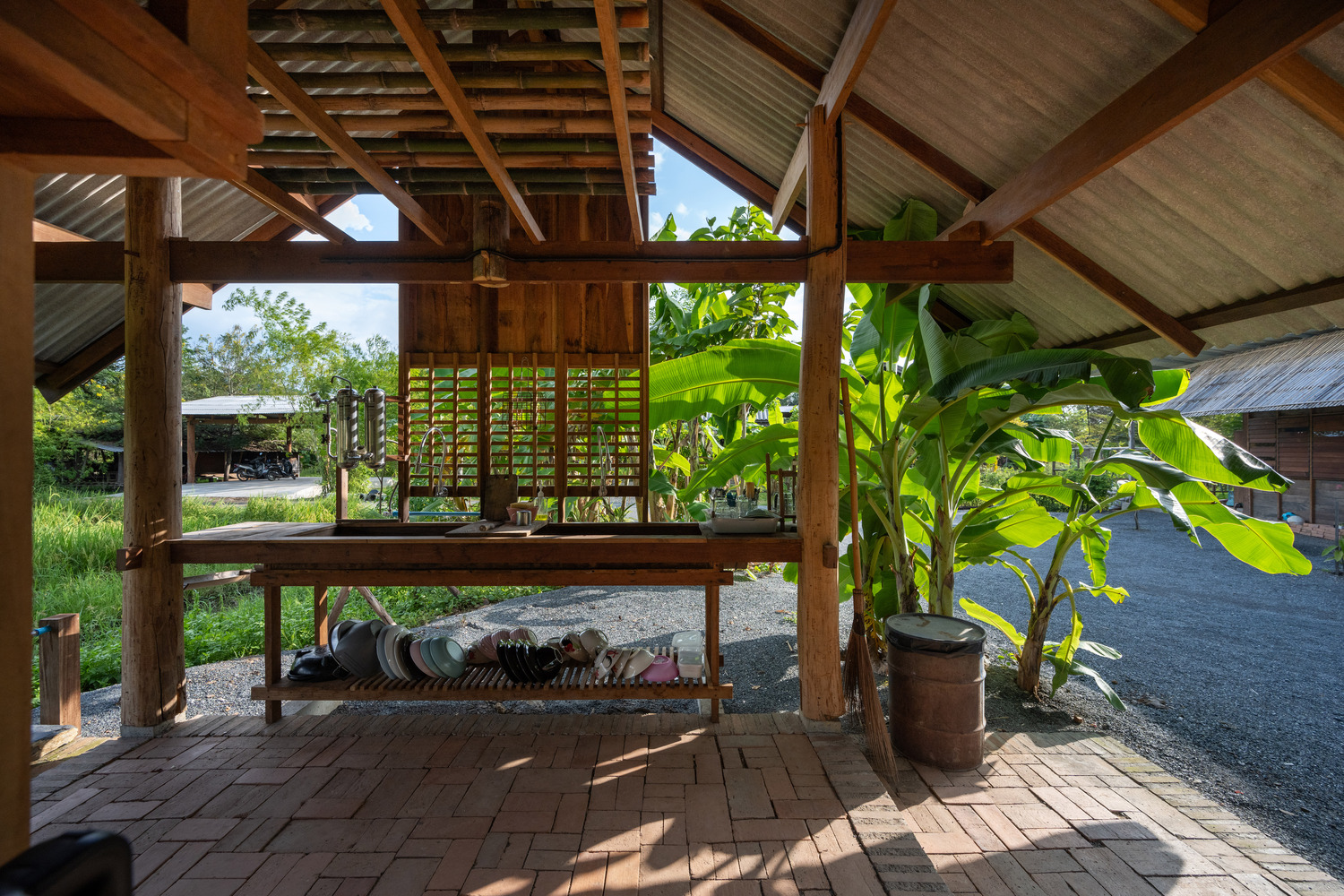 Open-air pavilion with timber frame and corrugated metal roof beside potted banana plants and rice fields