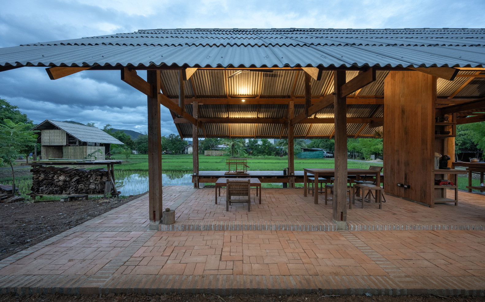 Open pavilion with timber columns and corrugated metal roof overlooking flooded rice fields at twilight