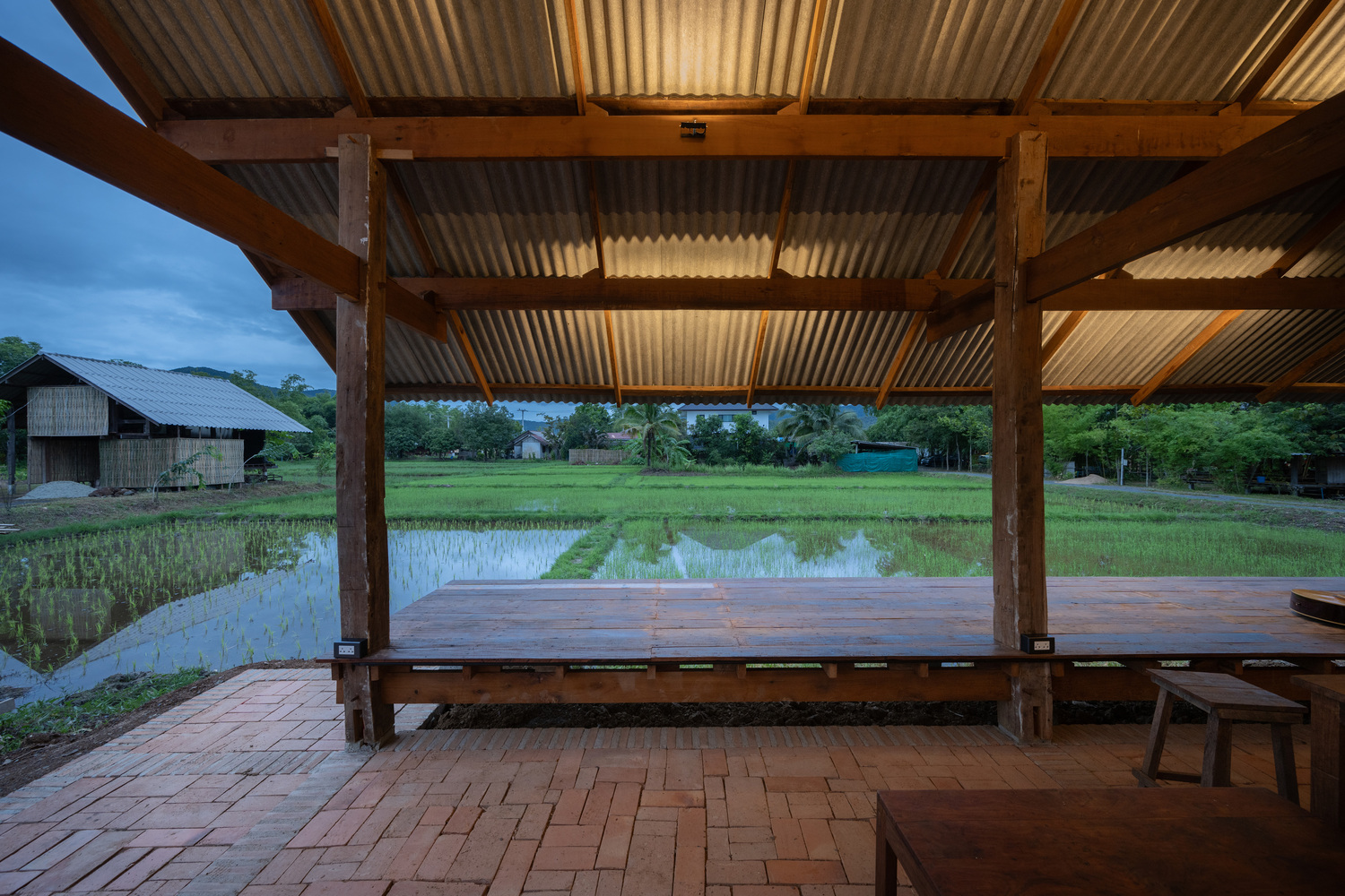 Covered terrace with timber columns, corrugated metal roof and brick paving overlooking rice paddies at dusk