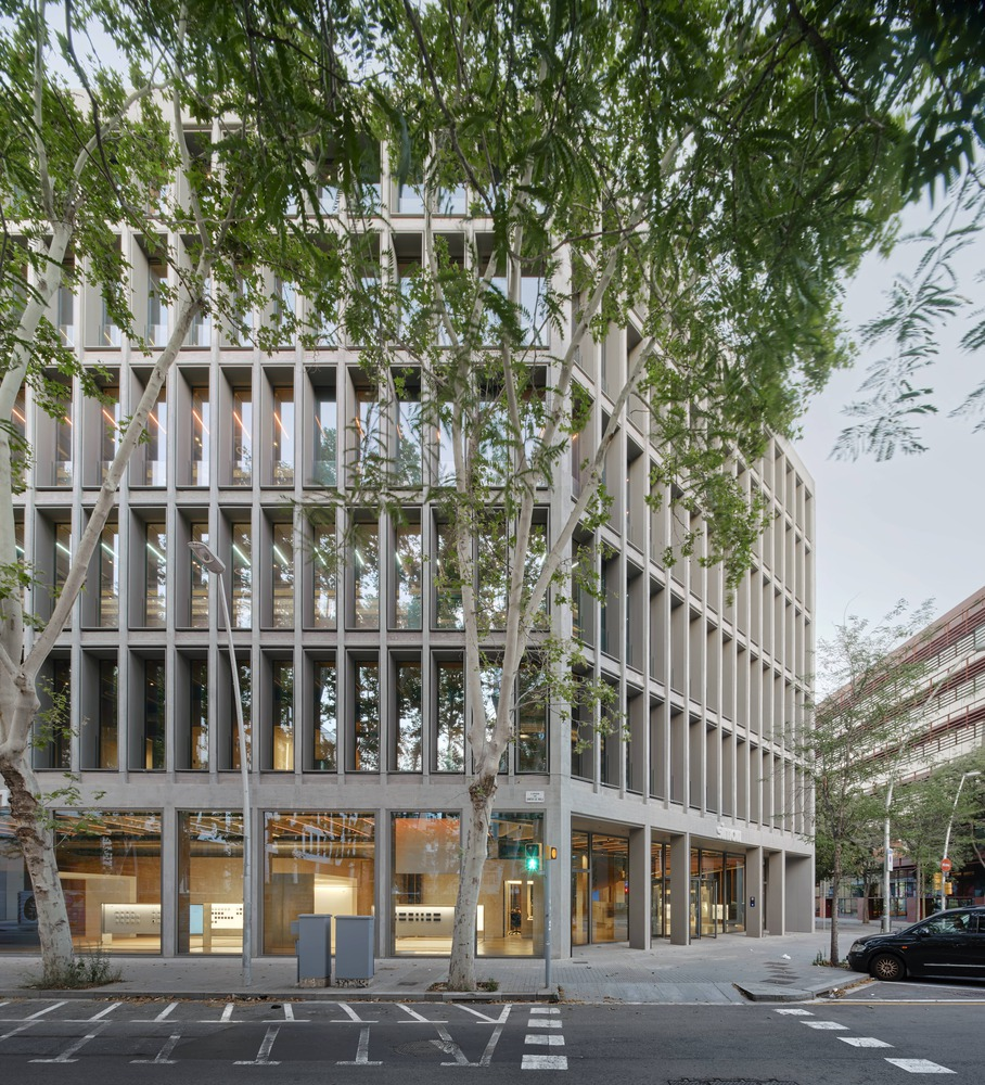 Full street elevation: six-storey concrete frame with vertical fins, glazed ground floor, trees along the pavement