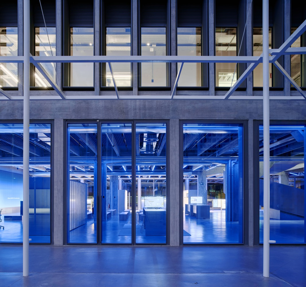Ground floor at night: blue-lit showroom space visible through full-height glass, steel structure behind