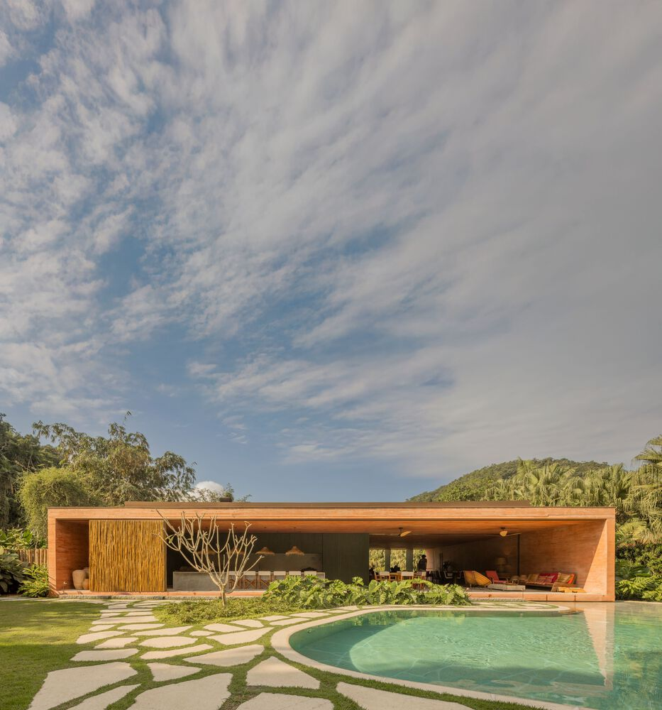 Long timber pavilion with deep overhang beside curving pool and flagstone path under cloudy sky