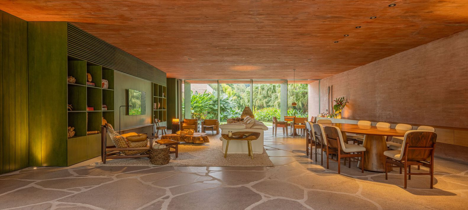 Open-plan living and dining space with timber ceiling, stone floor, and glass wall opening to garden