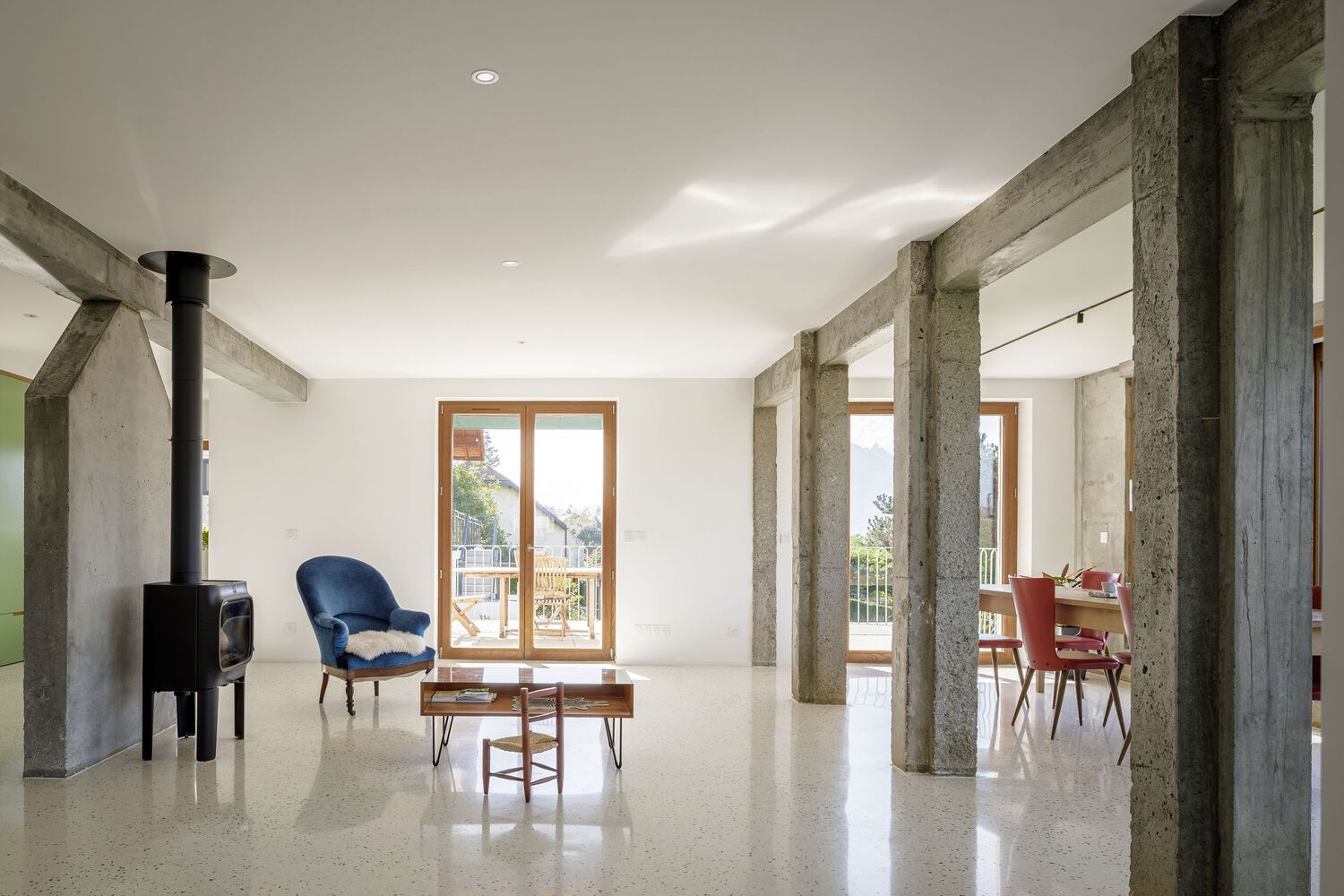 Open-plan living: exposed concrete columns and beam, terrazzo floor, wood stove, blue velvet chair, dining beyond