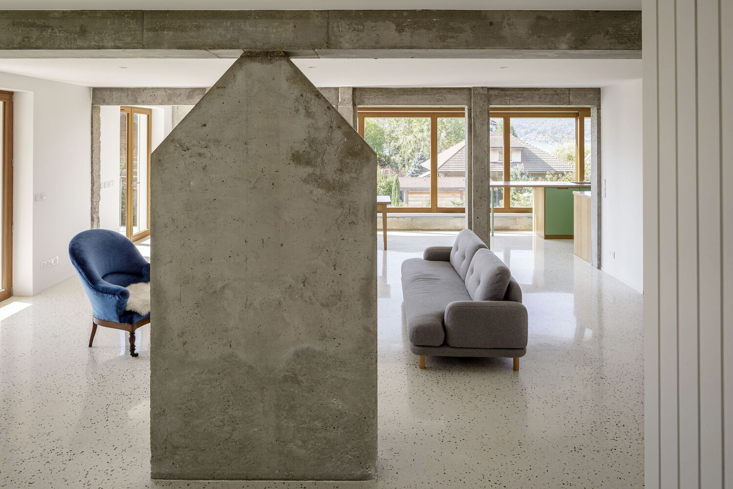 Living room from fireplace: house-shaped concrete chimney, grey sofa behind, timber doors, mountain light