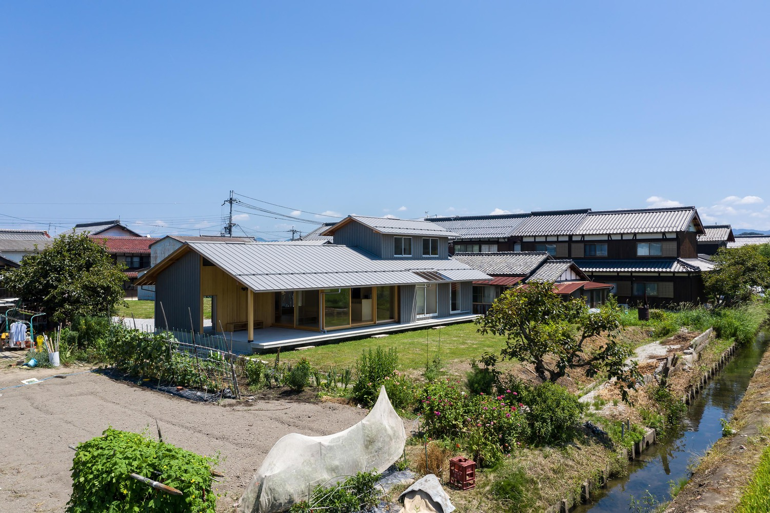 Village context: the house with its low metal roof sitting among traditional Japanese houses, stream, vegetable gardens