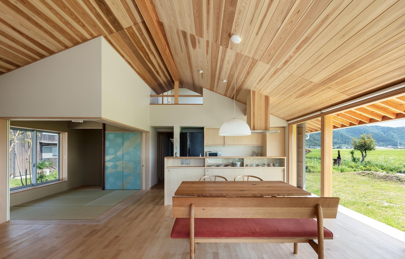 Kitchen and tatami: cedar ceiling, dining bench, kitchen island, blue-green sliding doors to the tatami room, mountains through glass