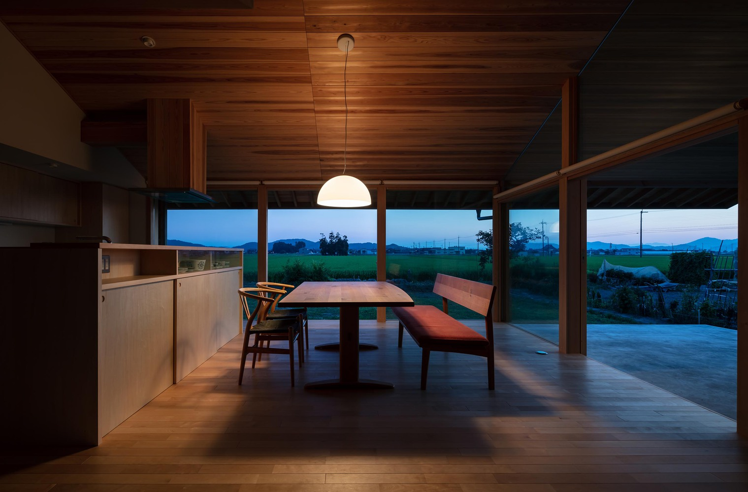 Dining at dusk: cedar interior glowing under pendant lamp, mountains silhouetted through full-width window, deep blue sky