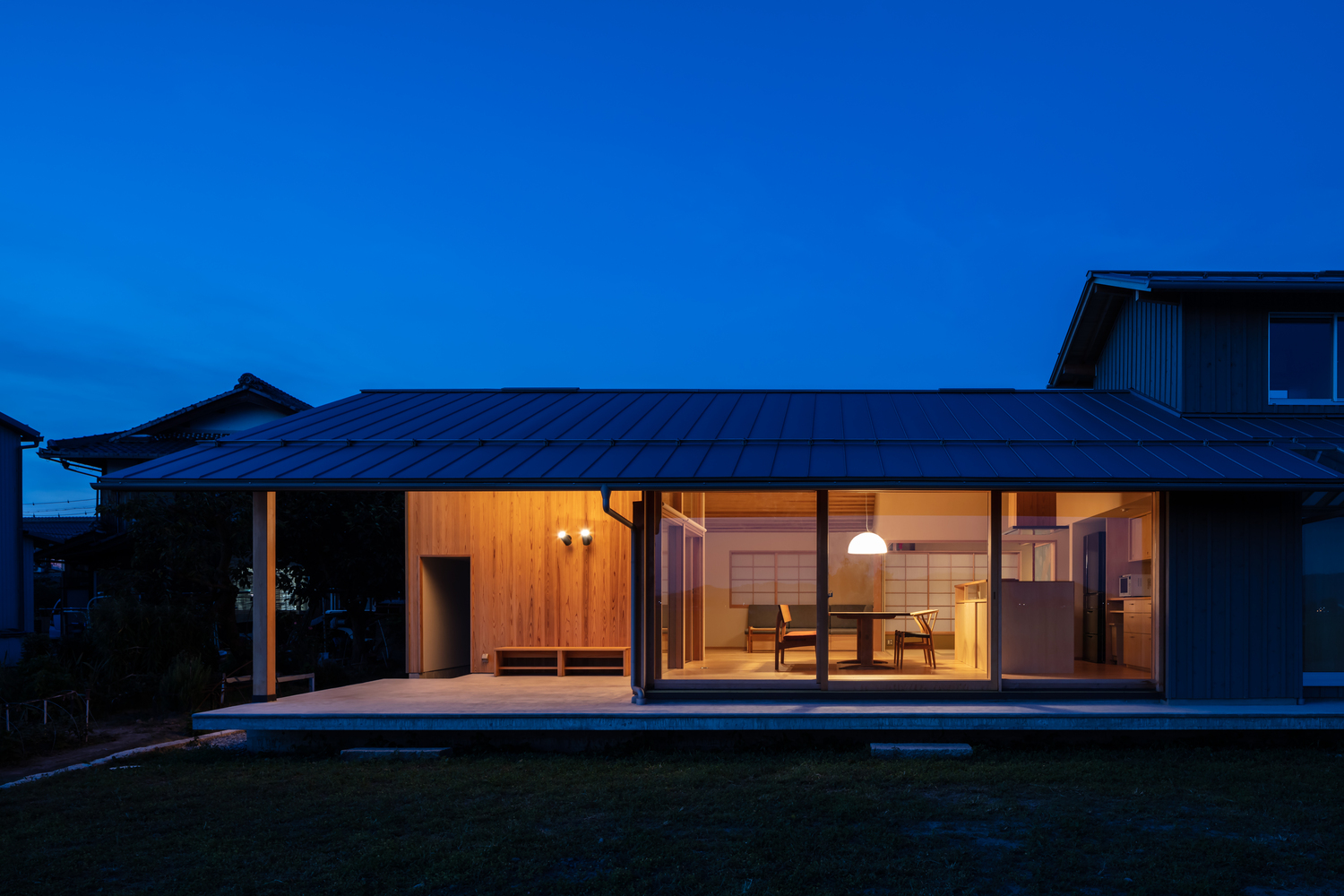 Night view from garden: the house as a glowing cedar box, kitchen and living room visible through glass, metal roof, dark sky