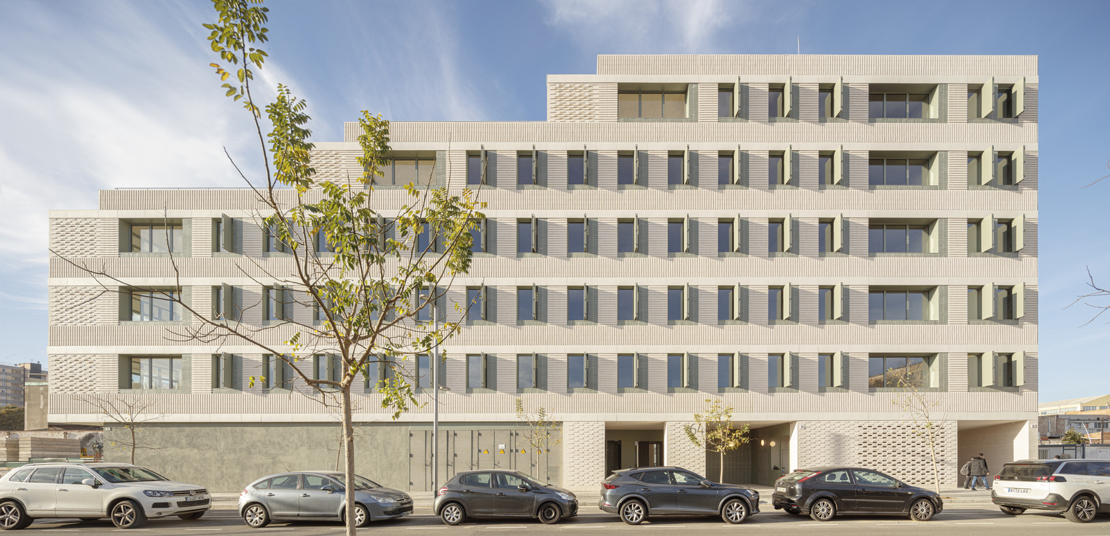 Street facade: six-storey cream brick building with stepped upper levels, perforated ceramic screen at ground floor, parked cars, young tree