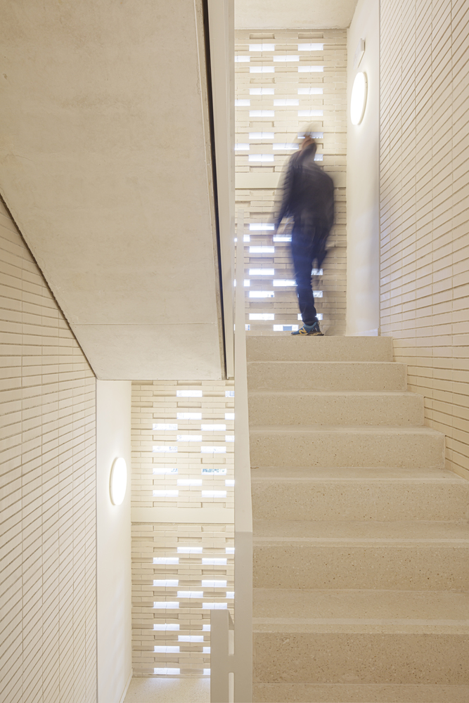 Stairwell: figure ascending concrete stairs, perforated ceramic screen casting patterned light on the walls, timber handrail