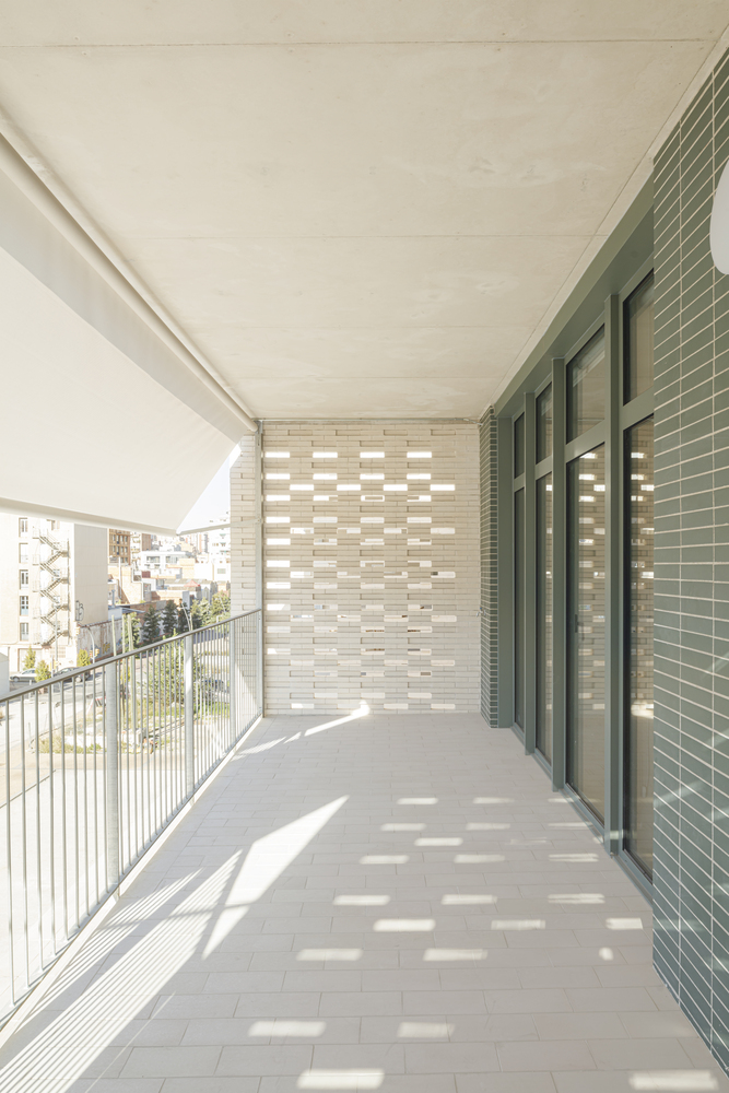 Balcony interior: deep terrace with perforated ceramic screen filtering sunlight, metal railing, concrete soffit, city rooftops beyond
