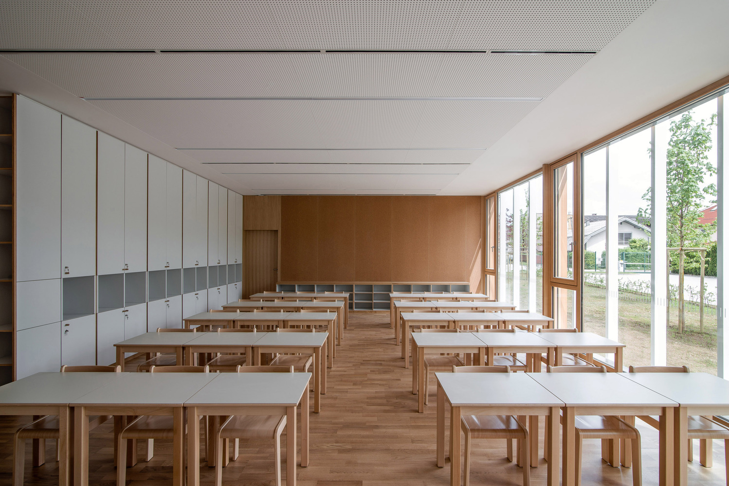 Classroom: timber desks and chairs, cork rear wall, white storage cabinets, full-height windows with garden view