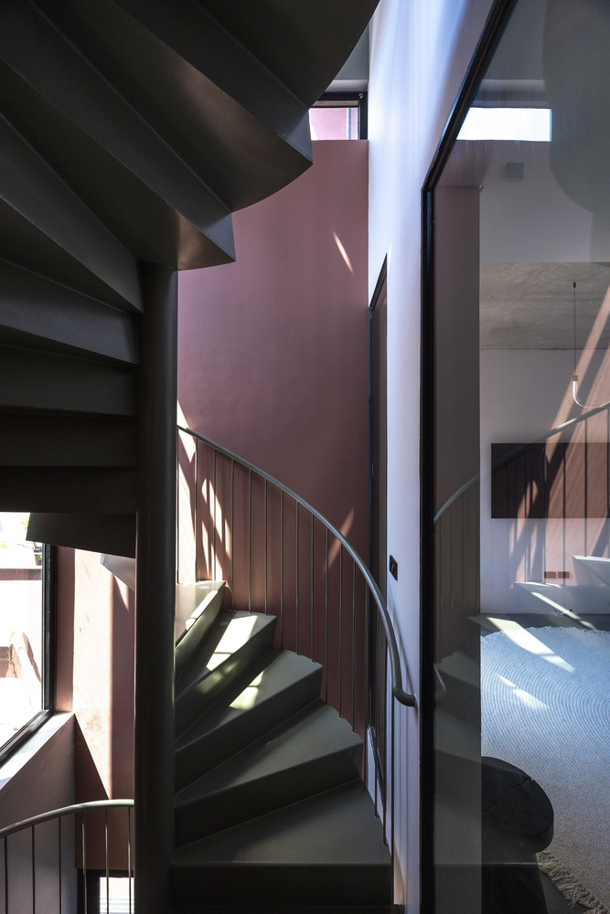 Steel spiral staircase with timber paneling ascending through a double-height space with filtered afternoon light