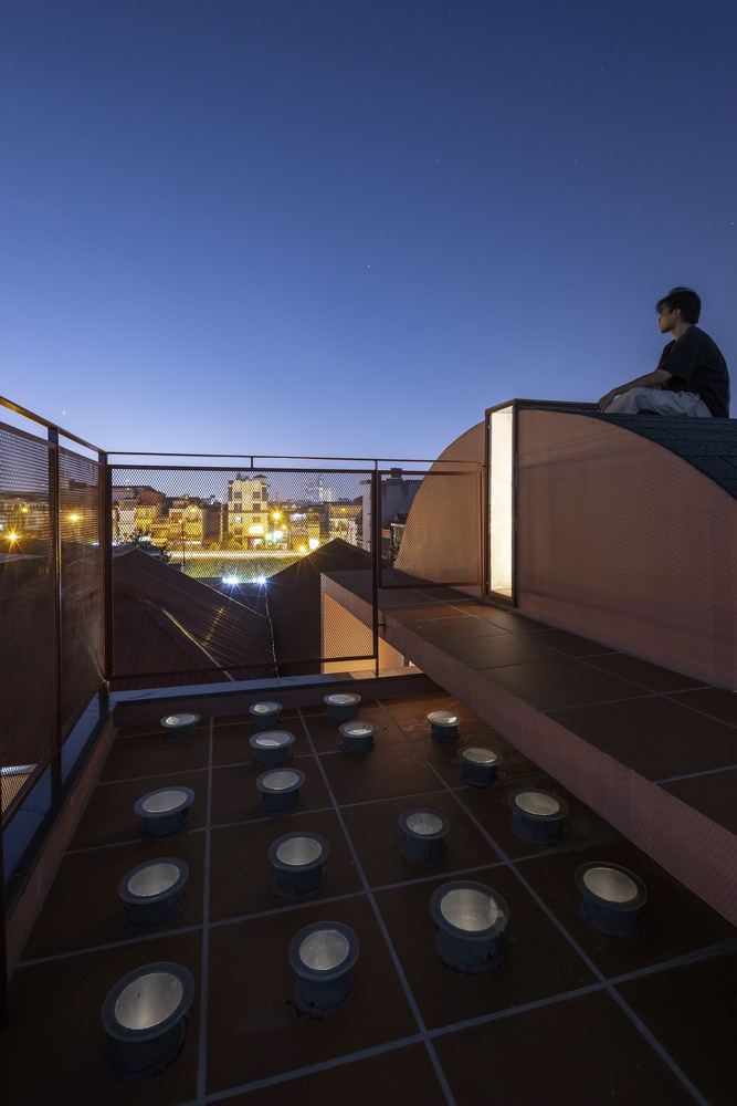 Rooftop terrace at twilight with person seated on curved illuminated volume and skylights set into tile floor
