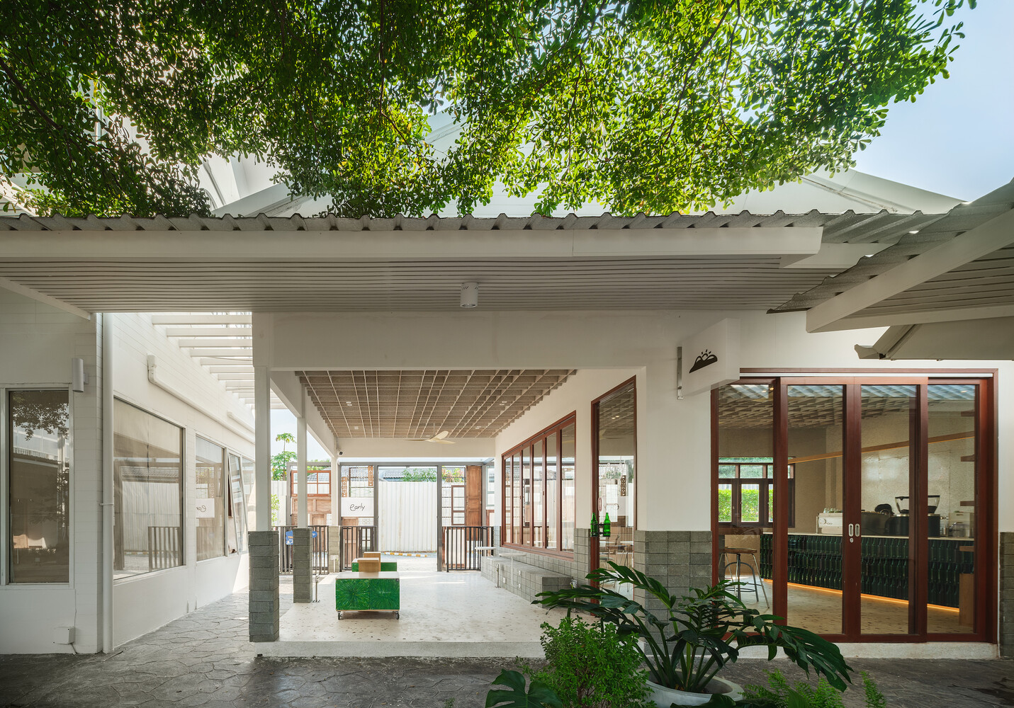 Semi-outdoor courtyard: white columns, reclaimed tile roof, tree canopy above, planting at ground level