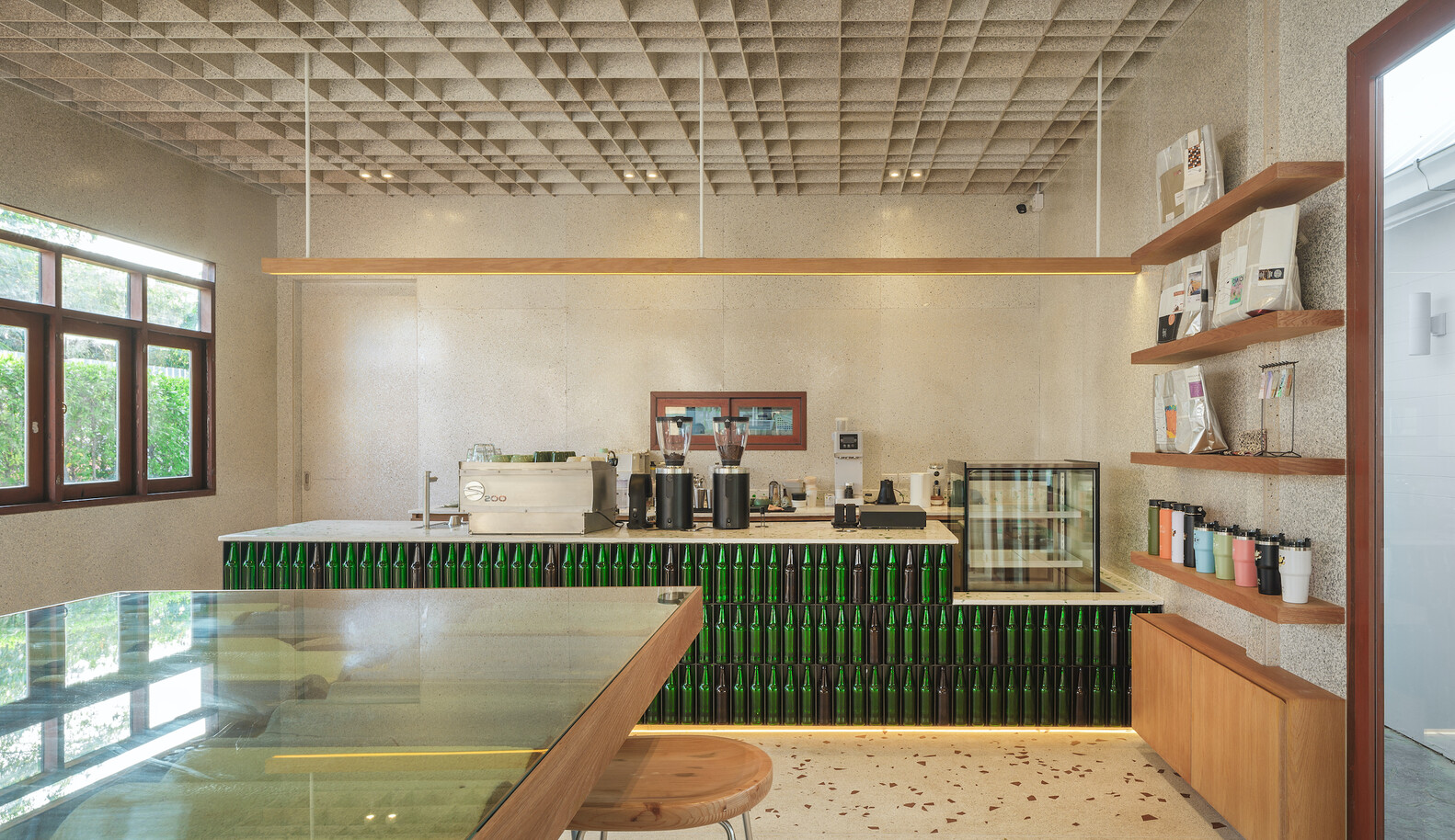 Counter from the side: green bottle bar, espresso machine, terrazzo surface, timber shelving unit
