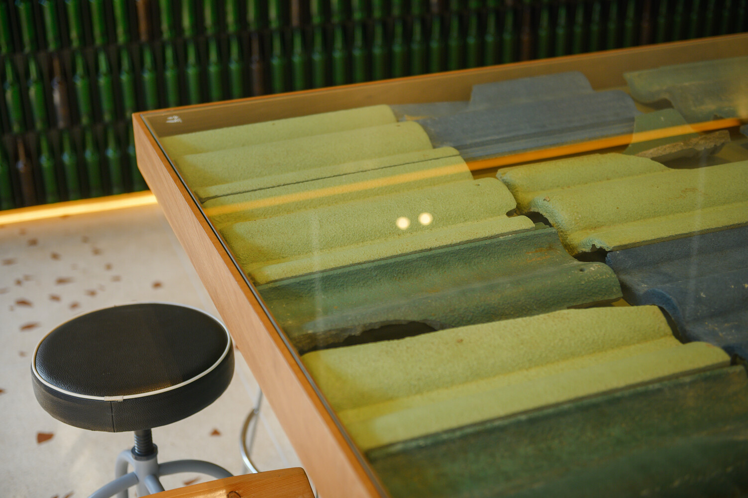 Counter table detail: reclaimed roof tiles under glass top, bar stool beside green bottles