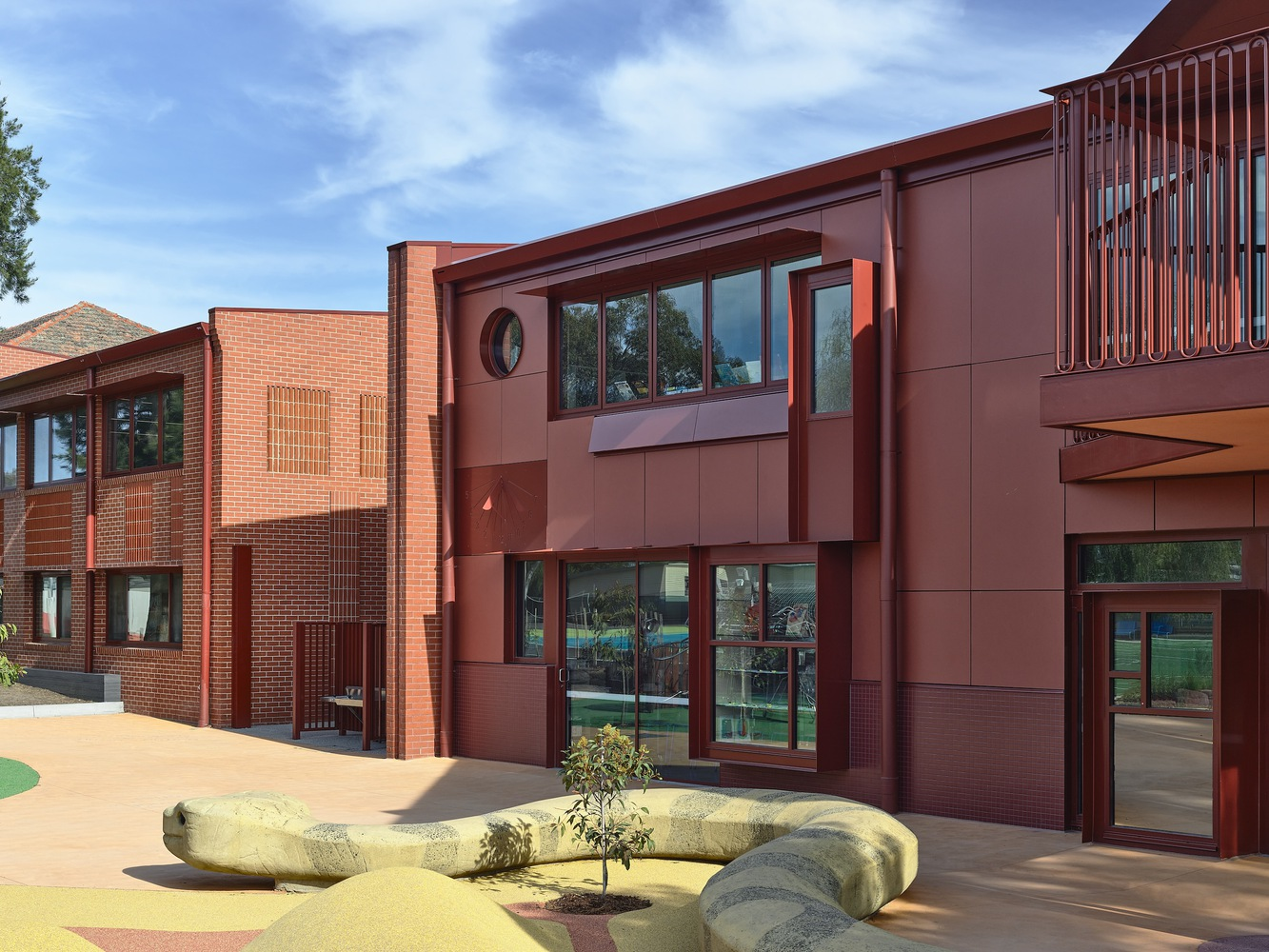 Courtyard view showing red brick and metal panel facades with a sculptural bench and young tree